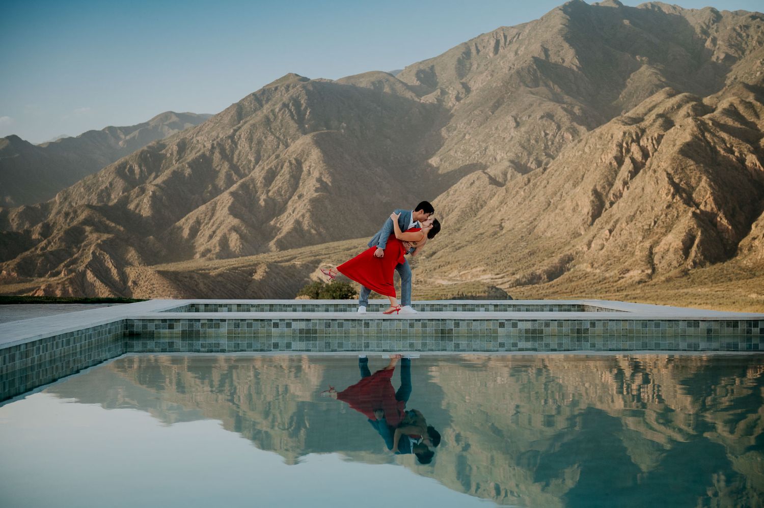 Couple sharing romantic dip kiss by reflective pool with stunning desert mountain backdrop.