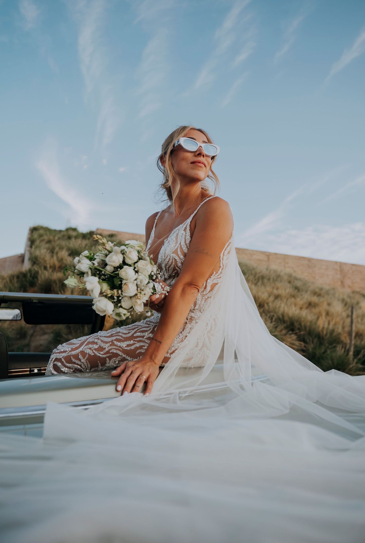 Bride in white lace gown and sunglasses holds bouquet atop a car with scenic golden hills at sunset.