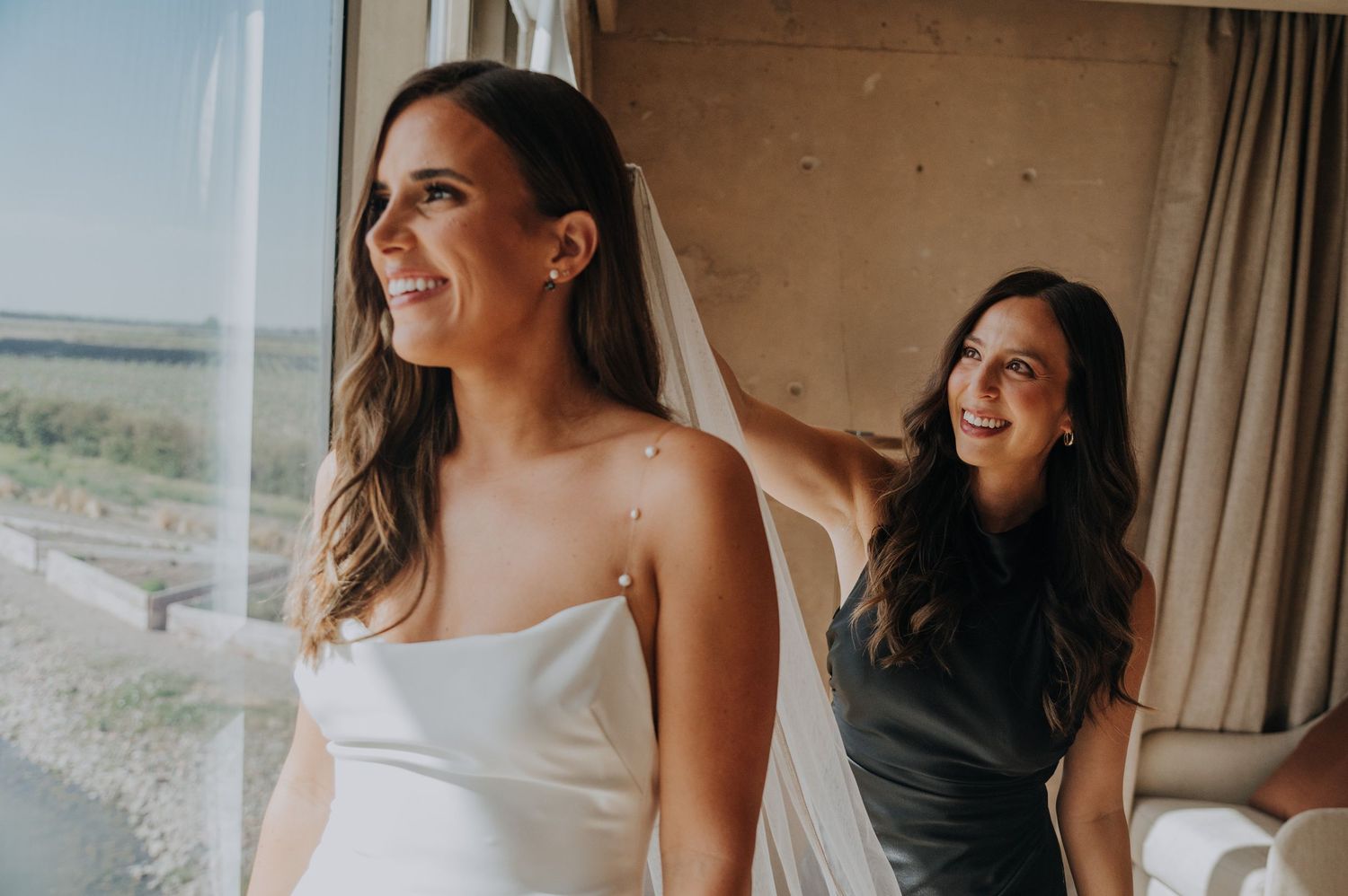 Bride in white gown smiles near window as bridesmaid in gray dress helps adjust her veil indoors.