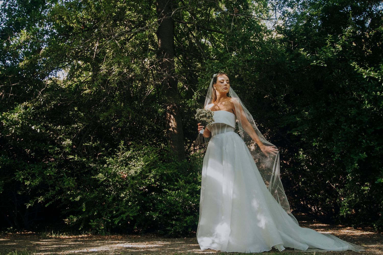 Bride in white strapless gown and veil poses gracefully among lush green trees in an outdoor wedding setting.
