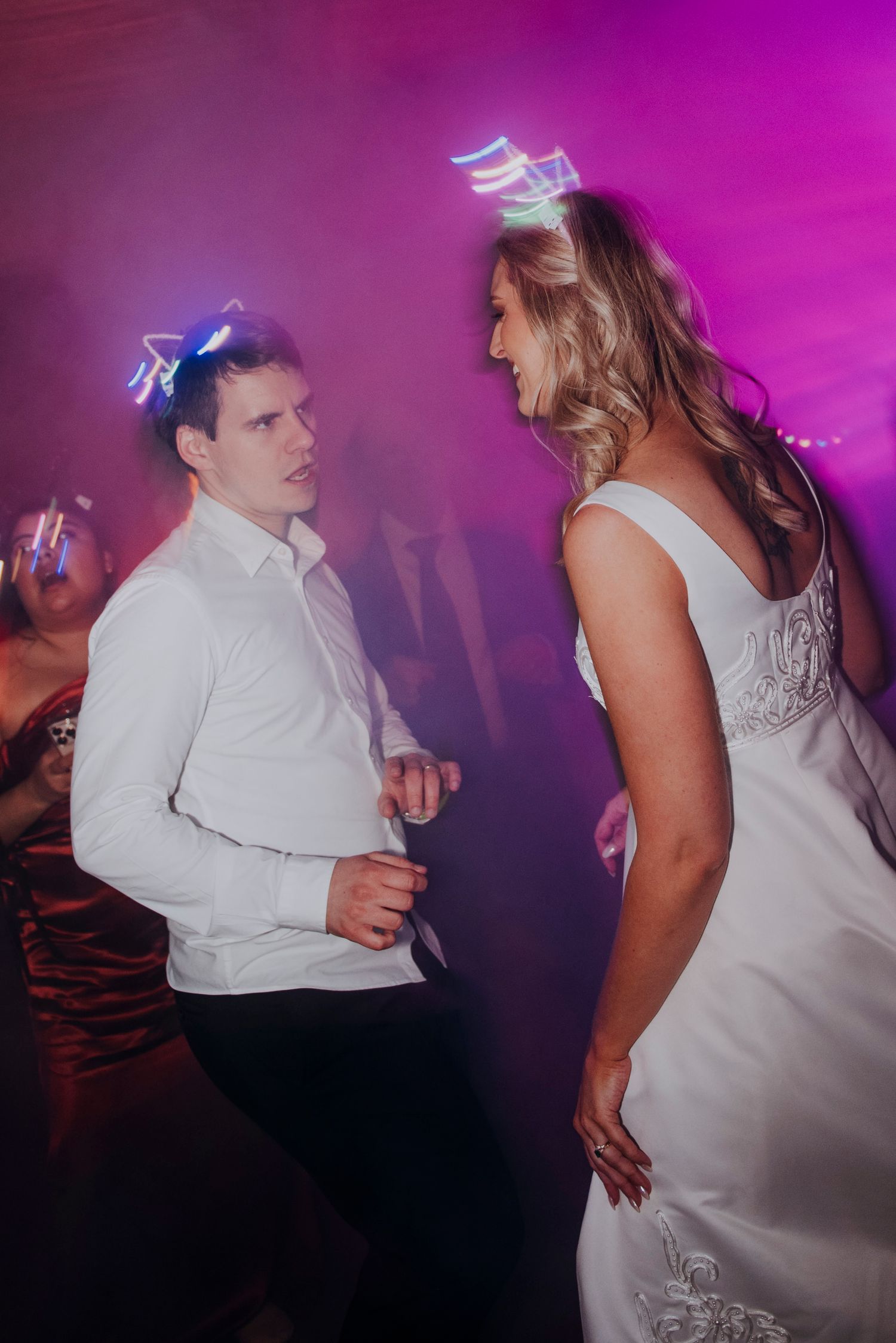 Bride and guest dance together under pink and purple lights at lively nighttime wedding reception.