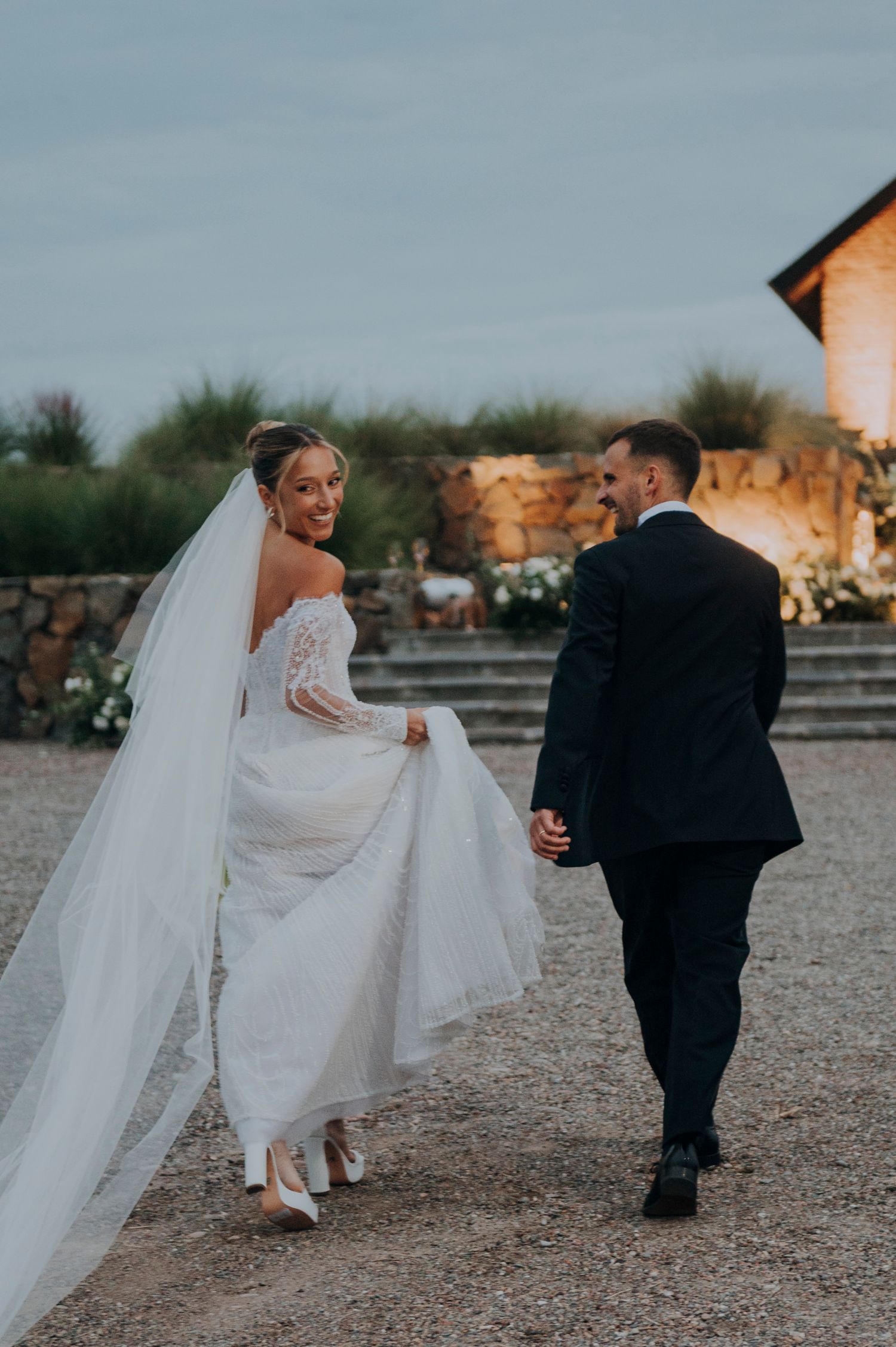 Bride in lace wedding gown with long veil walks hand-in-hand with groom at dusk.