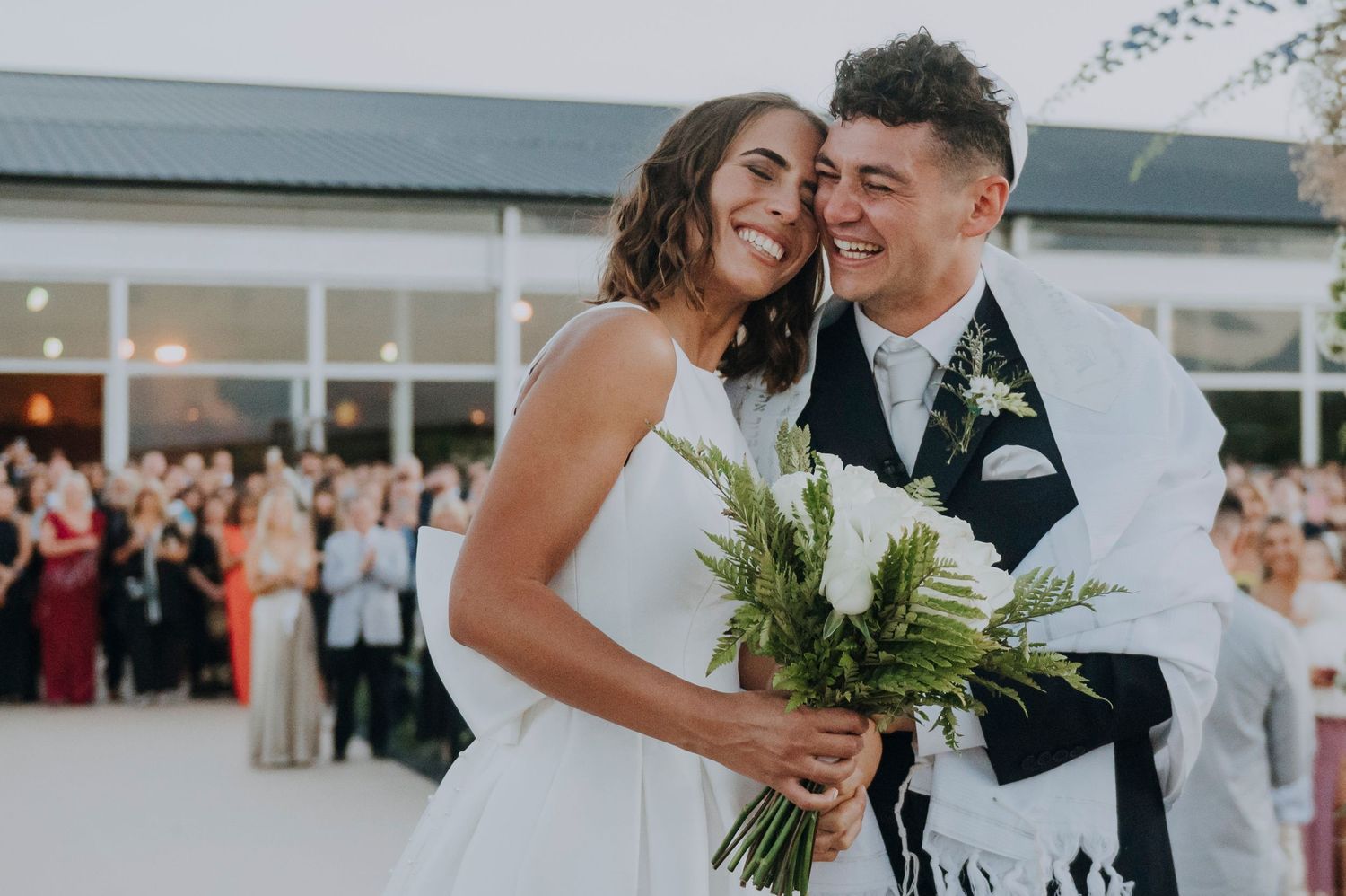 Joyful newlyweds smiling under a tallit at their outdoor wedding ceremony with guests celebrating in the background.