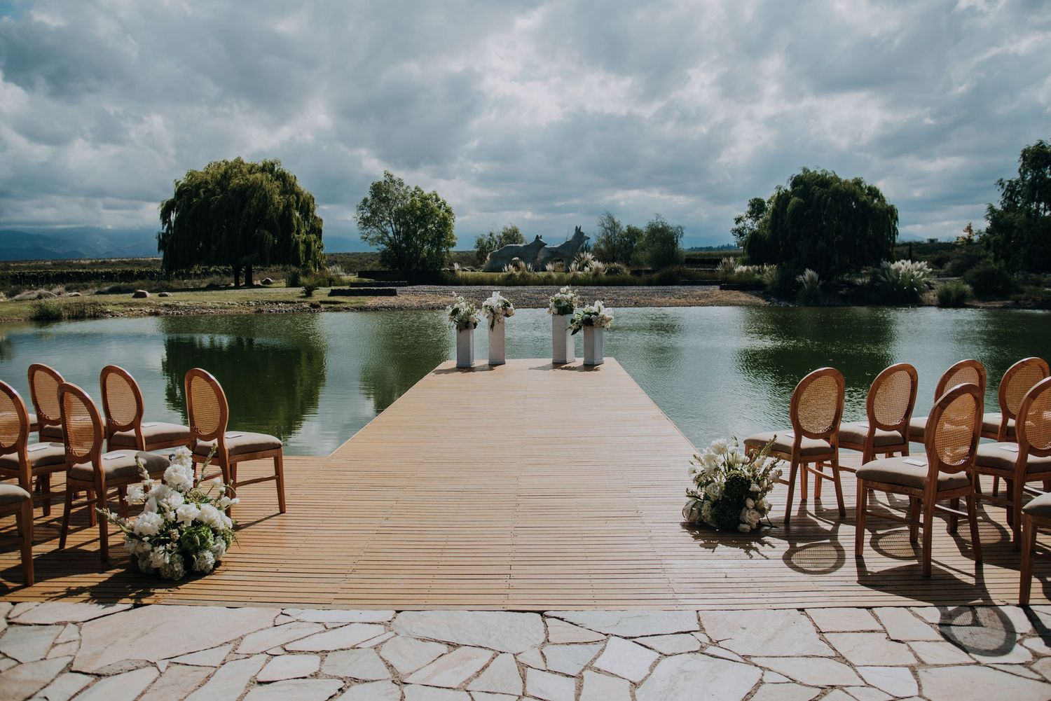 Outdoor wedding ceremony setup on wooden dock over lake with rattan chairs and floral arrangements under cloudy sky.