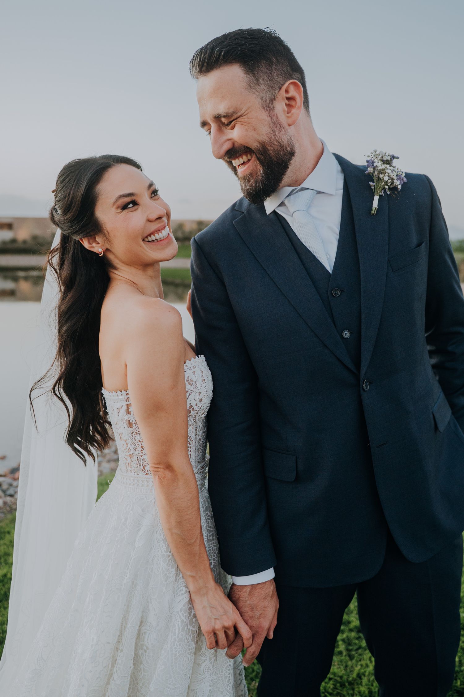 Smiling bride and groom holding hands outdoors, bride in strapless lace gown, groom in navy suit with boutonniere.