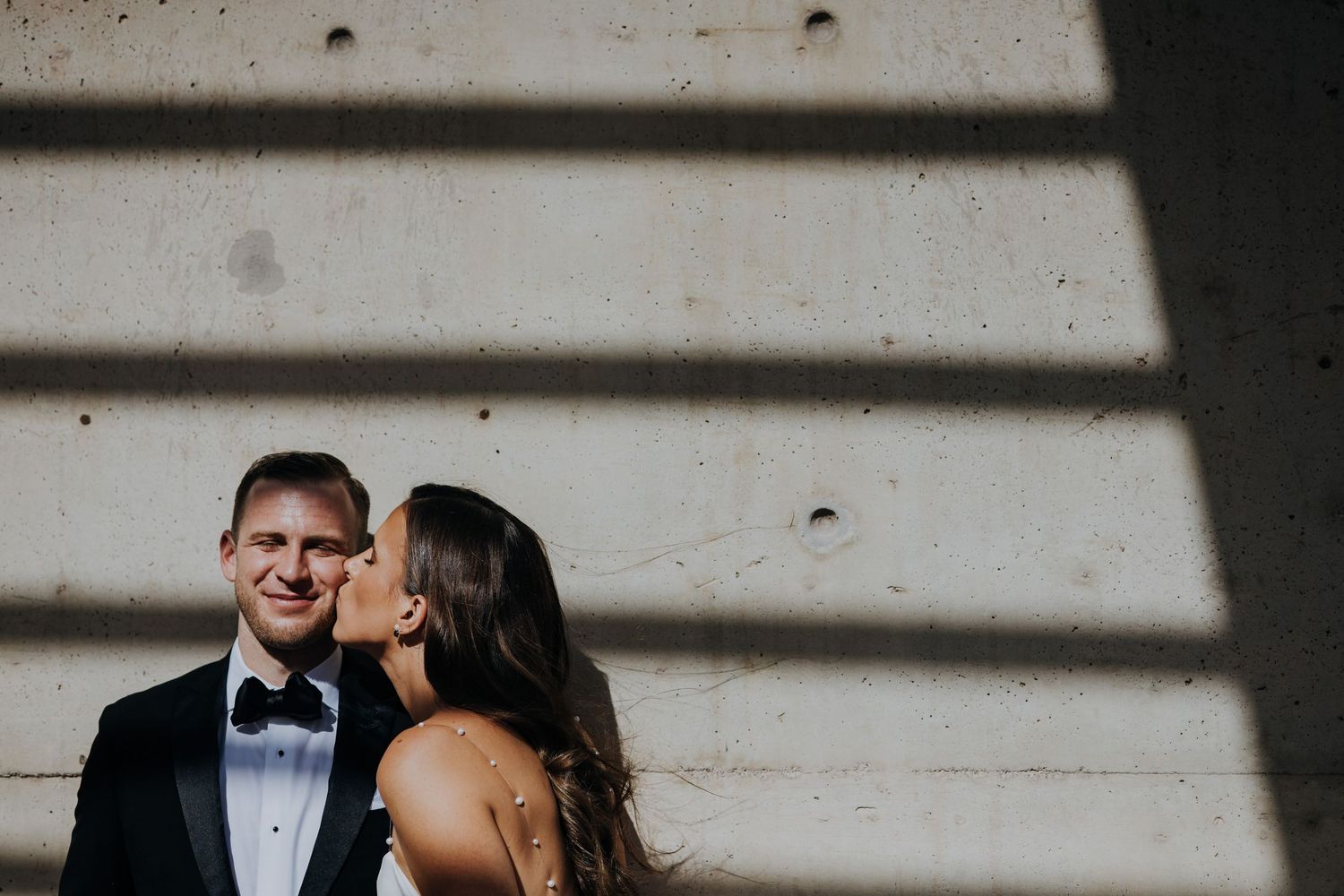 Bride kissing groom on the cheek as he smiles against a concrete wall with shadow stripes.