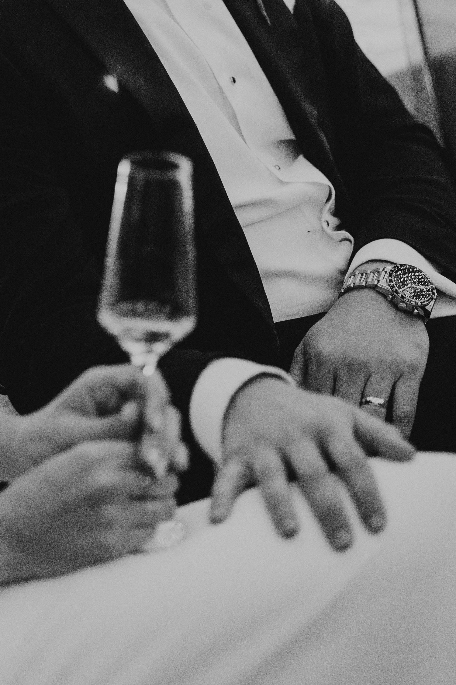 Black and white close-up of groom's hand with watch beside champagne flute at wedding reception.