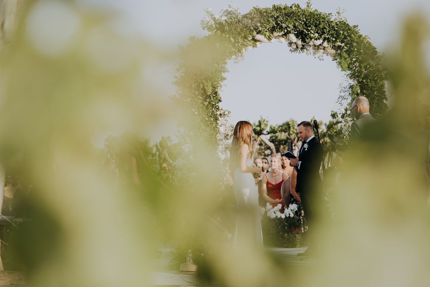 Couple exchanges vows under a circular greenery arch at an outdoor wedding ceremony.