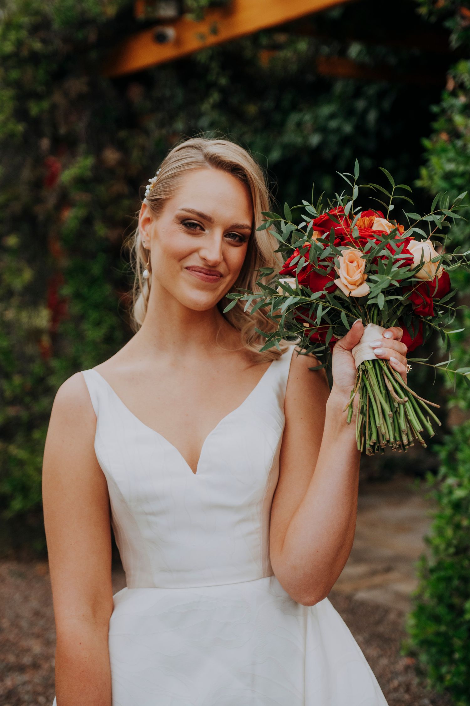 Elegant bride in white V-neck gown holding vibrant red and pink bridal bouquet outdoors.