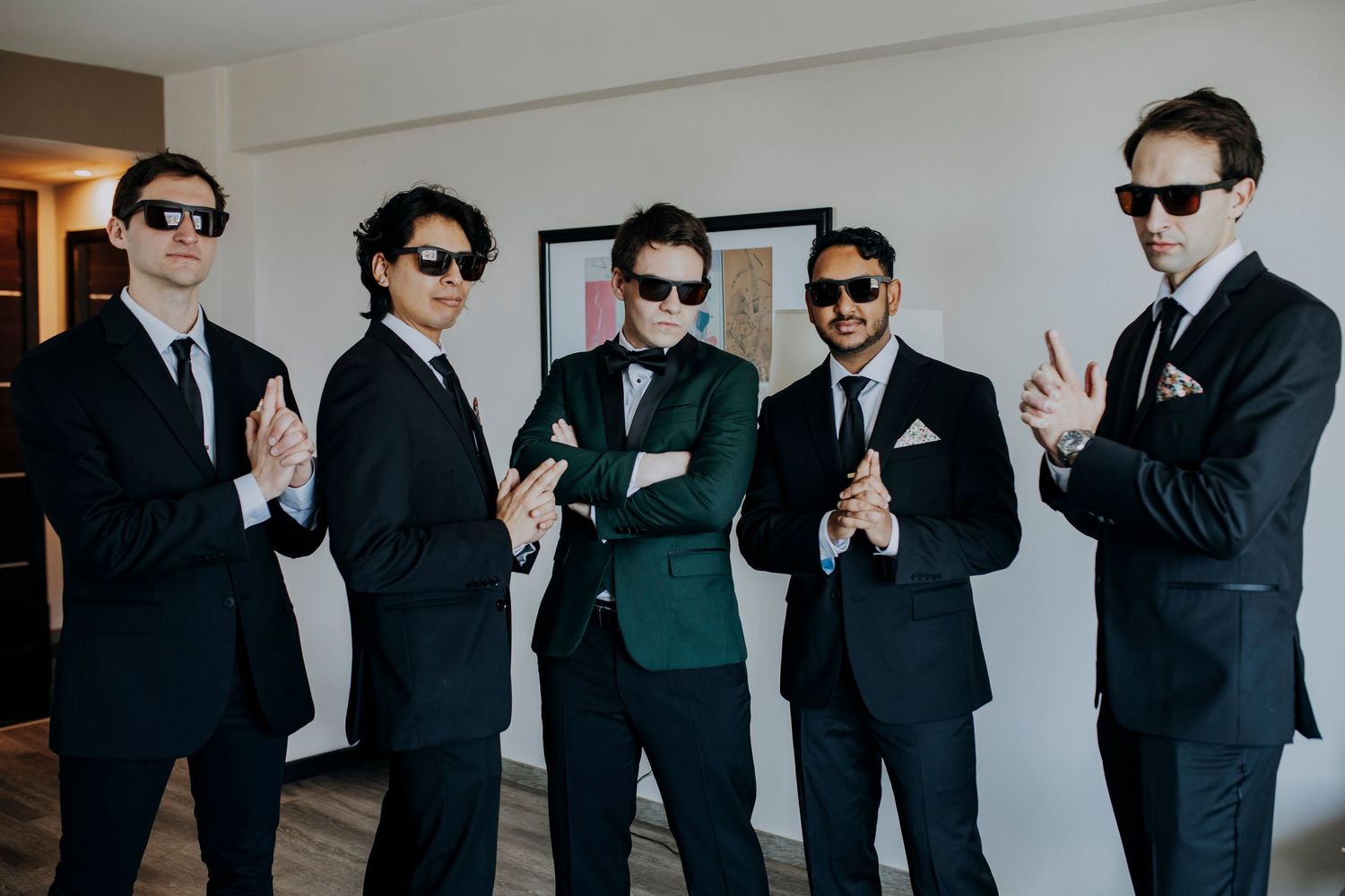 Five men in black suits and sunglasses pose confidently indoors, with one in a green tie standing out as the groom.
