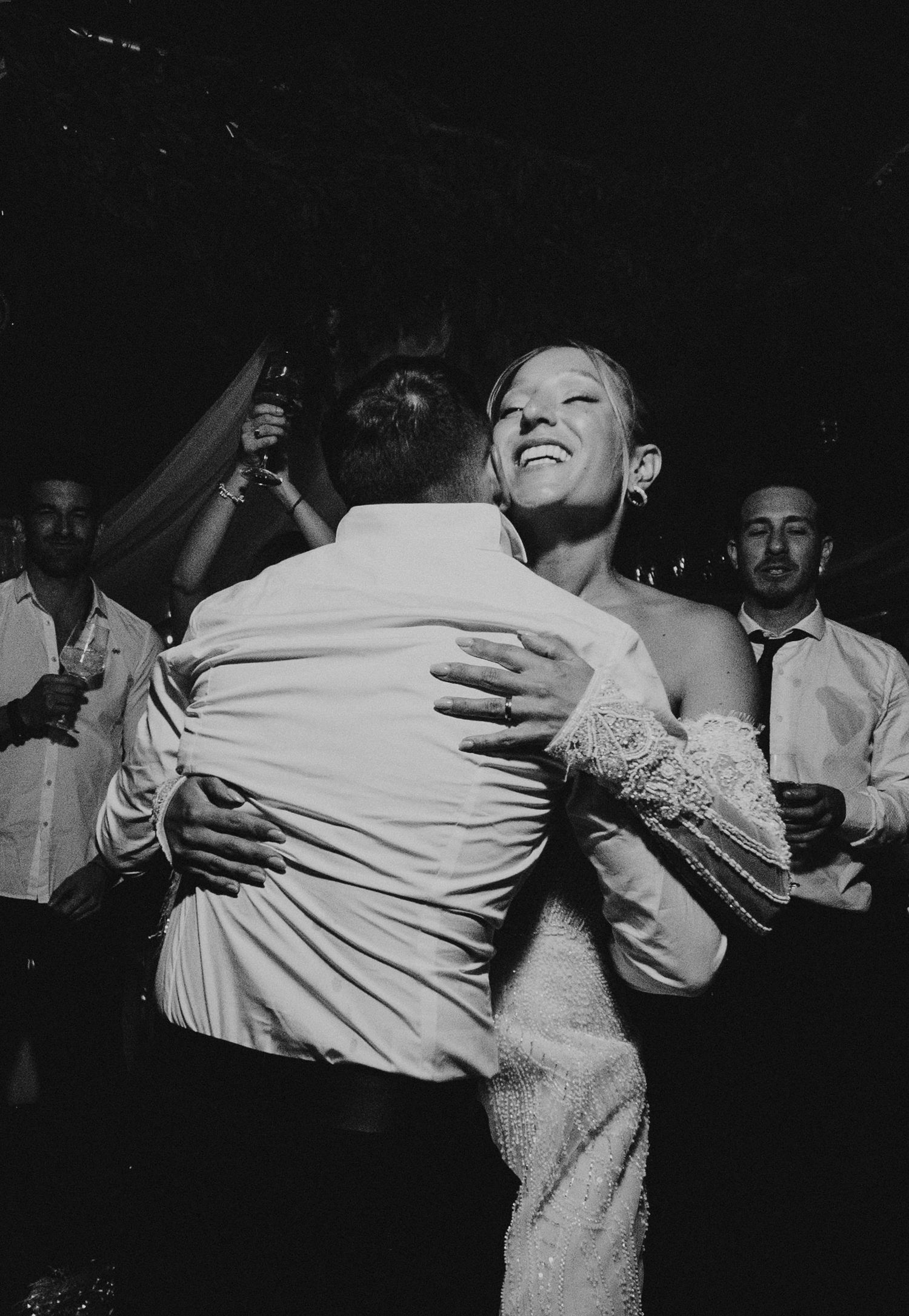 Black and white photo of bride and groom embracing on the dance floor surrounded by wedding guests.