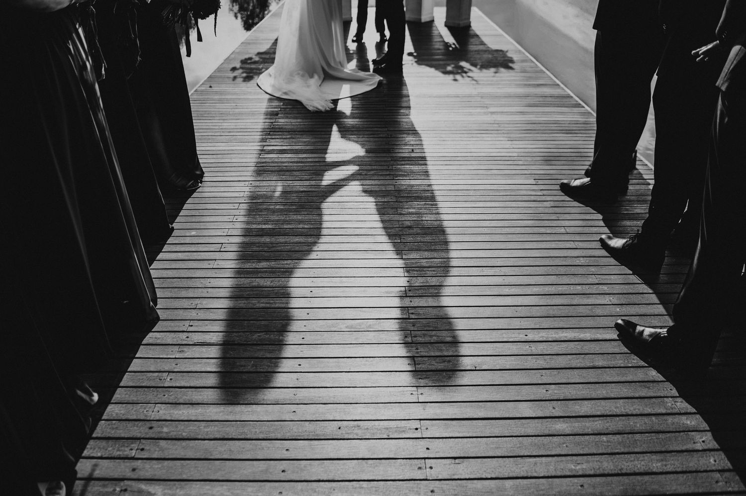 Black and white photo of wedding couple shadows cast on wooden deck surrounded by guests.