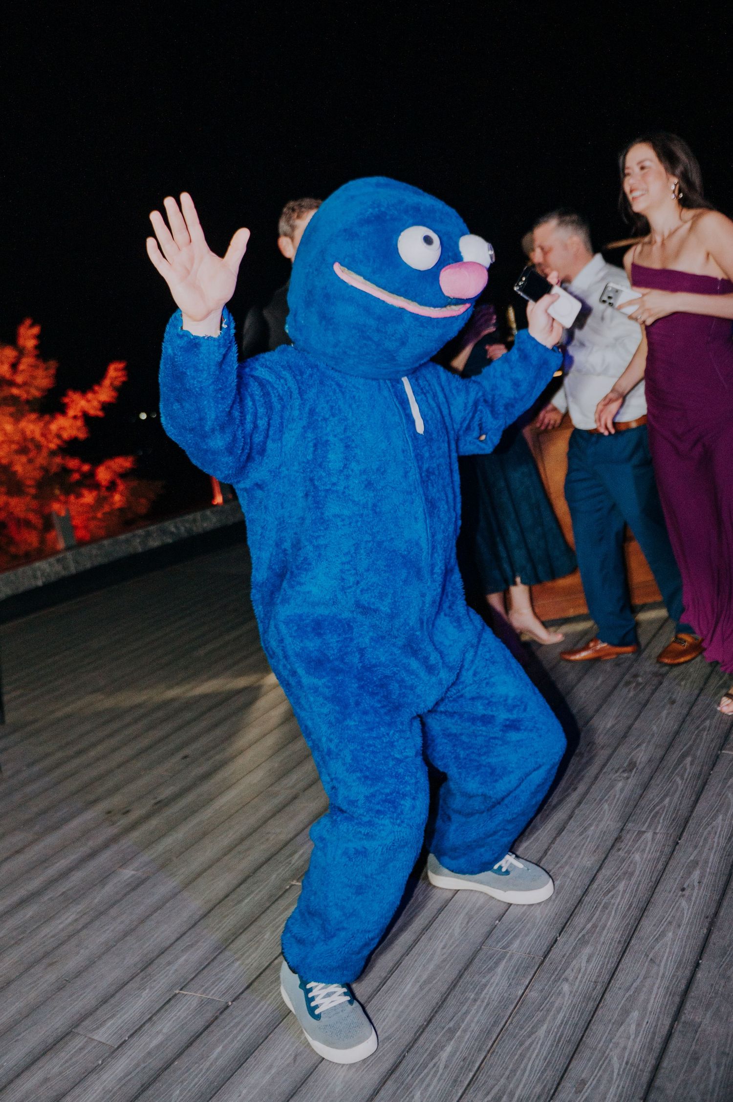 A person in a blue monster costume dances on an outdoor deck at a nighttime wedding reception.