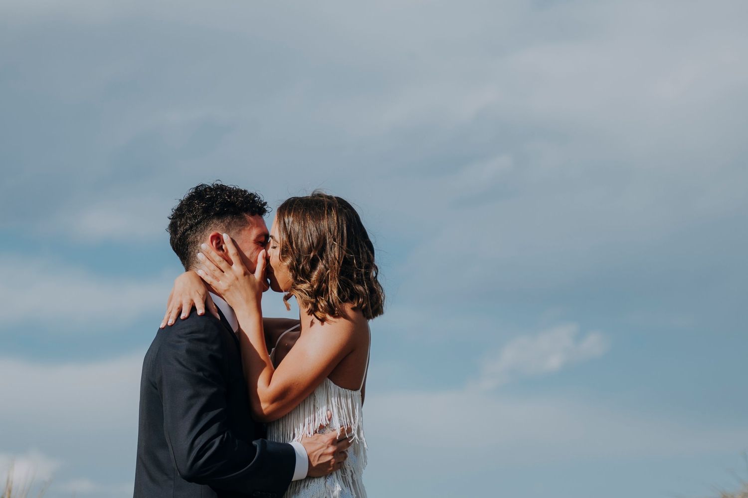 Couple sharing a romantic kiss outdoors against a dramatic blue cloudy sky, bride in white dress and groom in dark suit.