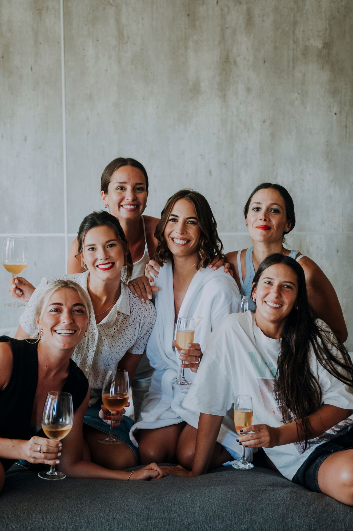 Bride in white robe celebrates with five bridesmaids holding champagne glasses during wedding prep.