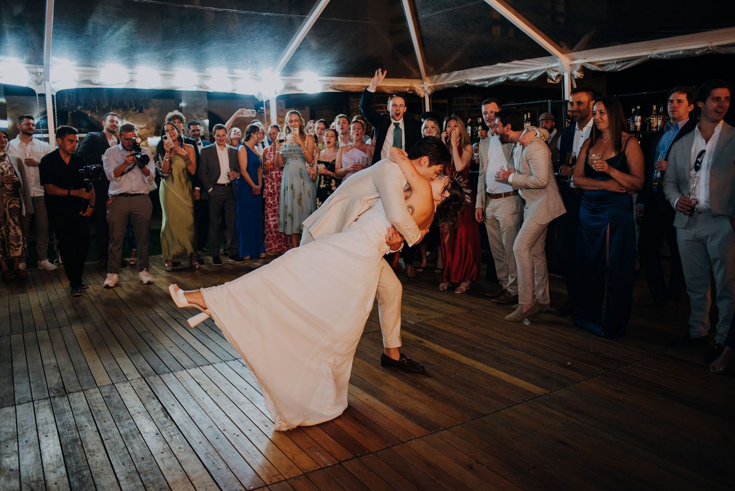 Groom dips bride dramatically on dance floor surrounded by cheering guests at wedding reception.