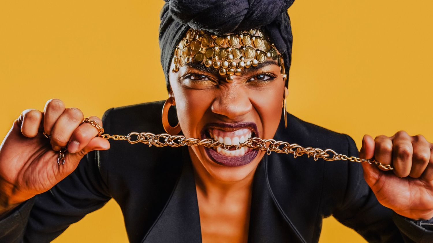 Bold woman in gold headwrap biting gold chain necklace against vibrant yellow background.