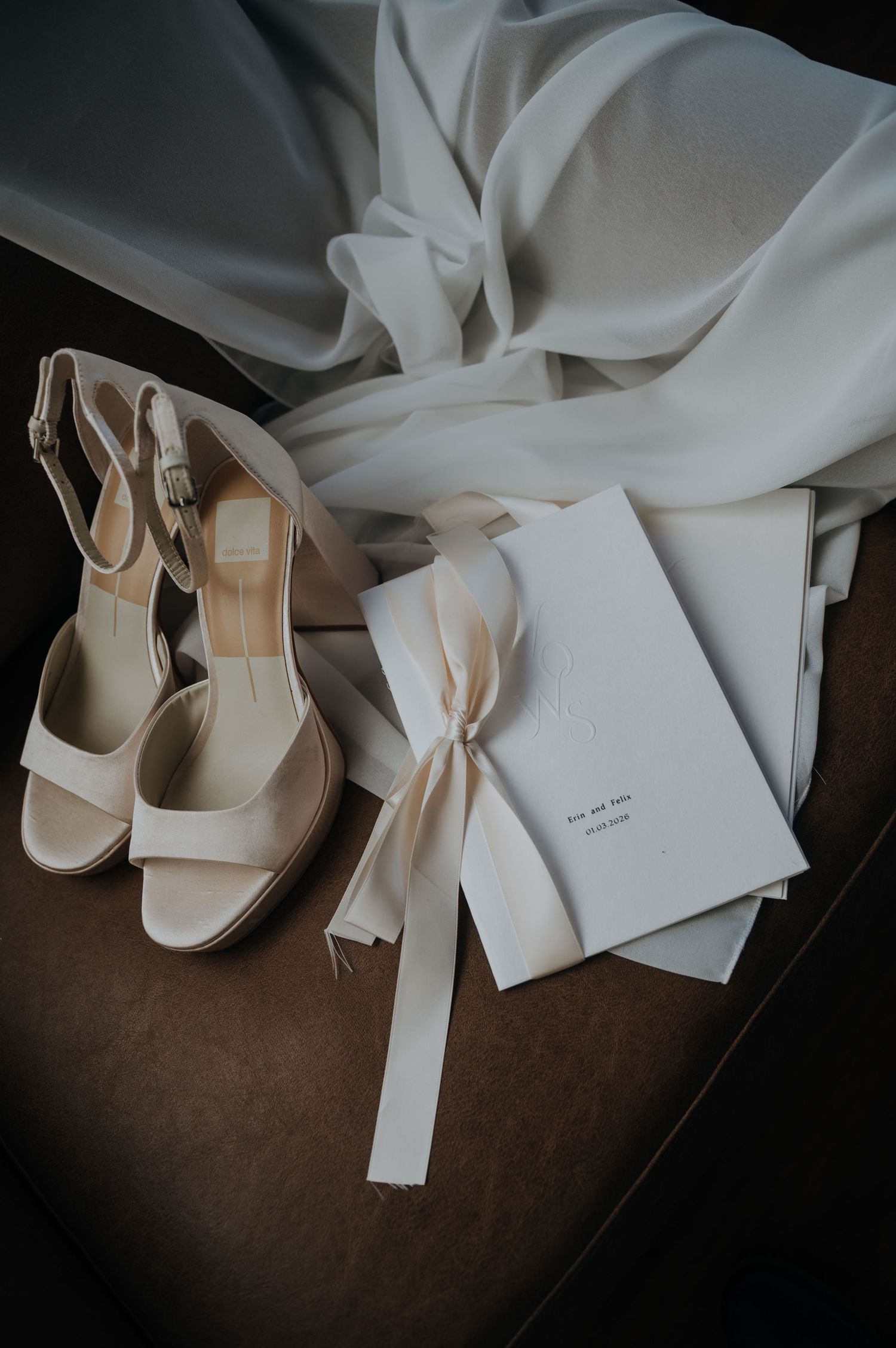 Elegant bridal details featuring ivory heeled sandals, a ribbon-tied wedding booklet, and flowing white veil on a brown chair.