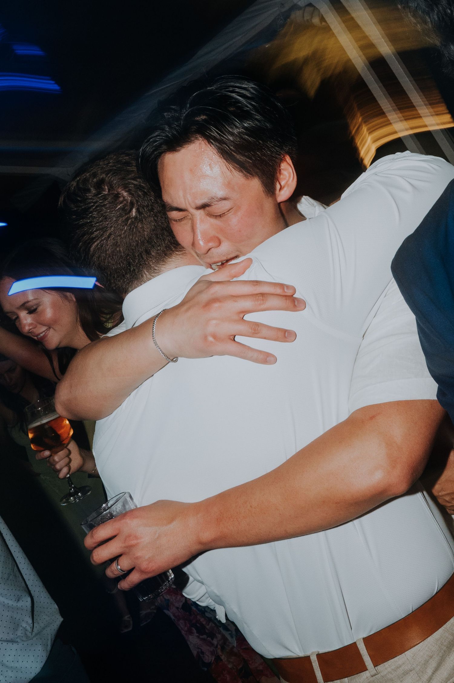 Two men sharing a warm hug at a party, one wearing a white shirt and bracelet holding a drink.