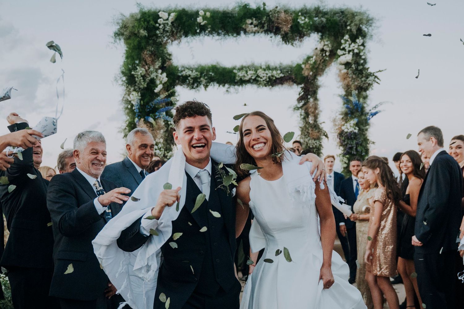 Joyful newlyweds walk down aisle as guests toss confetti beneath floral chuppah at outdoor wedding.