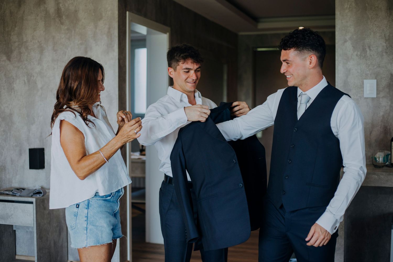 Groom being helped into navy suit jacket by groomsman and bridesmaid before wedding ceremony.