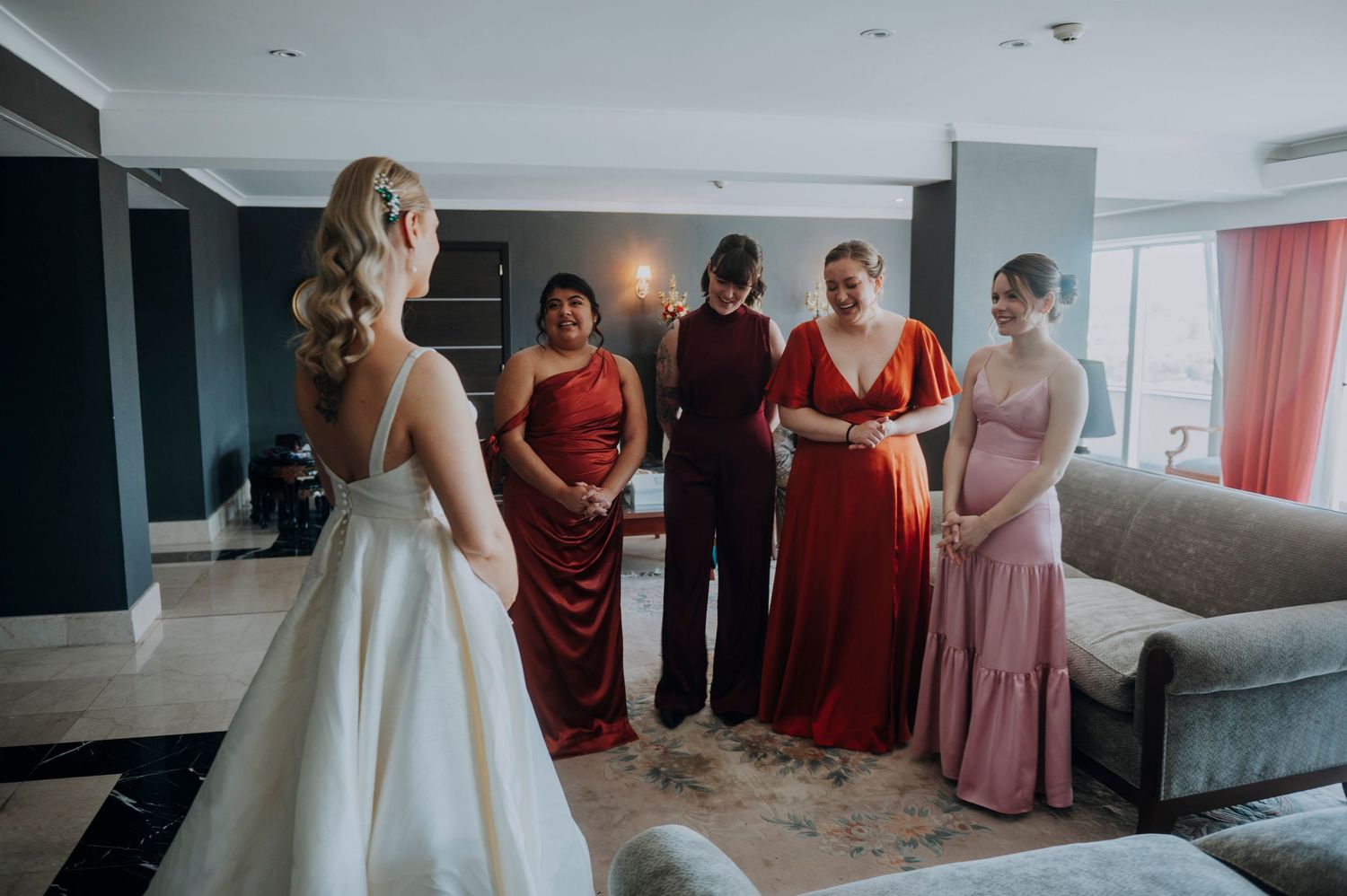 Bride in white gown reveals dress to bridesmaids in red and pink dresses in elegant hotel suite.