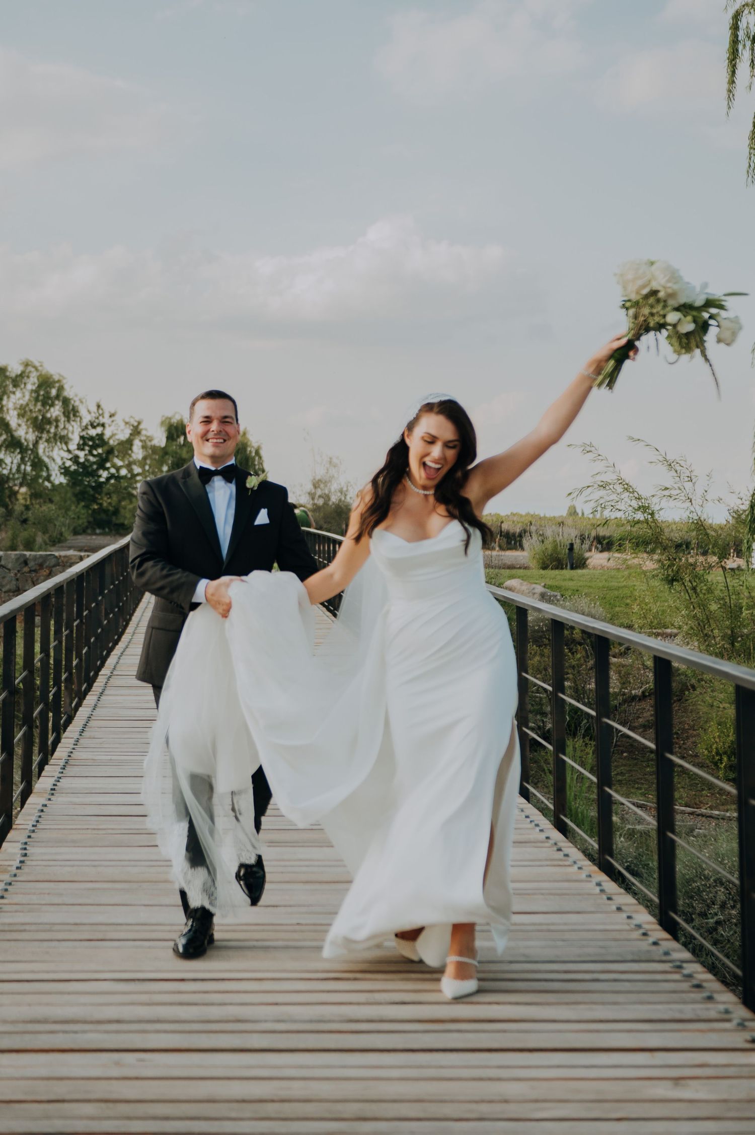 Joyful newlyweds laugh and run across a wooden bridge, bride raising bouquet in celebration after wedding.