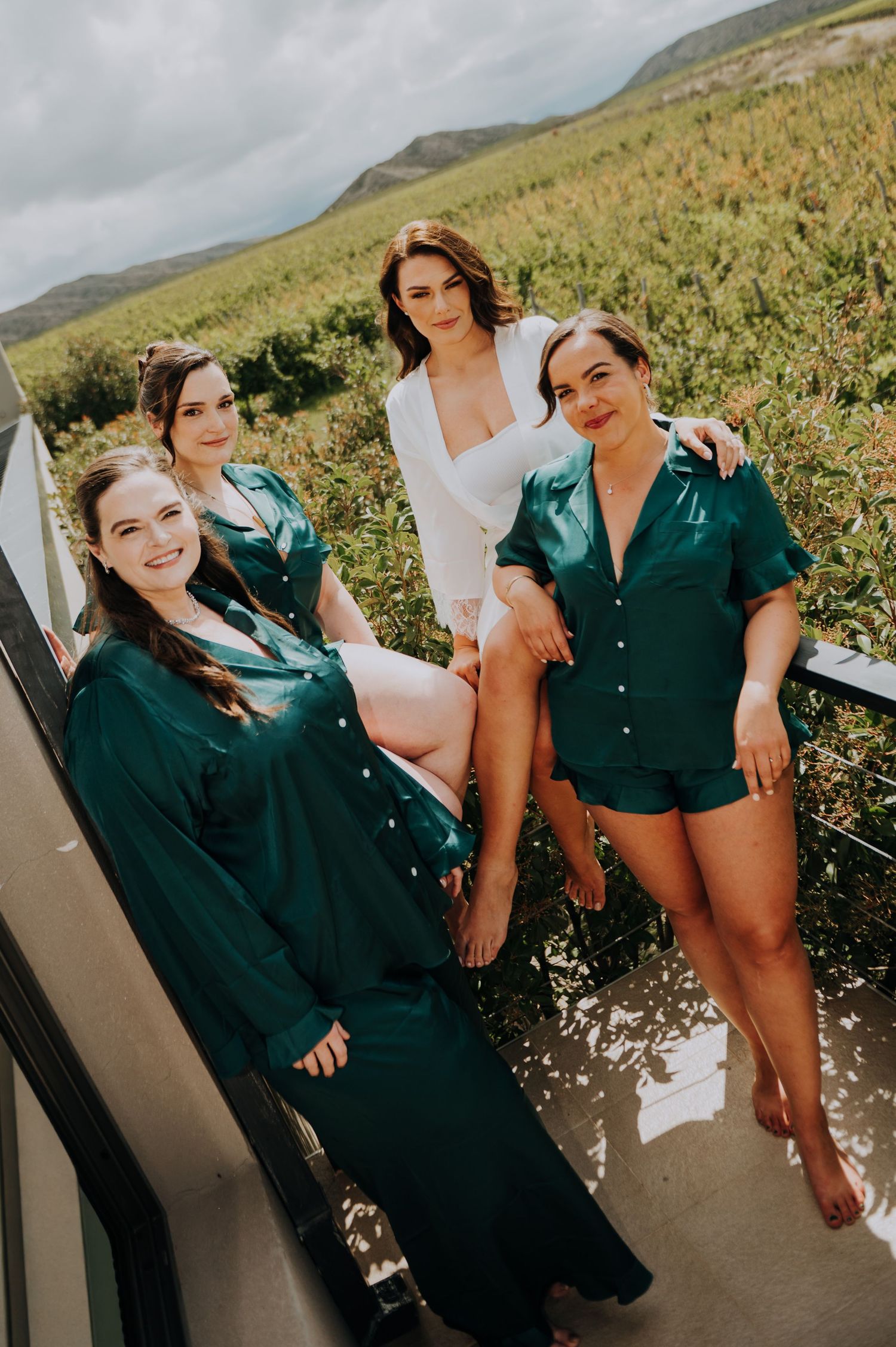 Bride in white with three bridesmaids in emerald green satin robes posing outdoors.