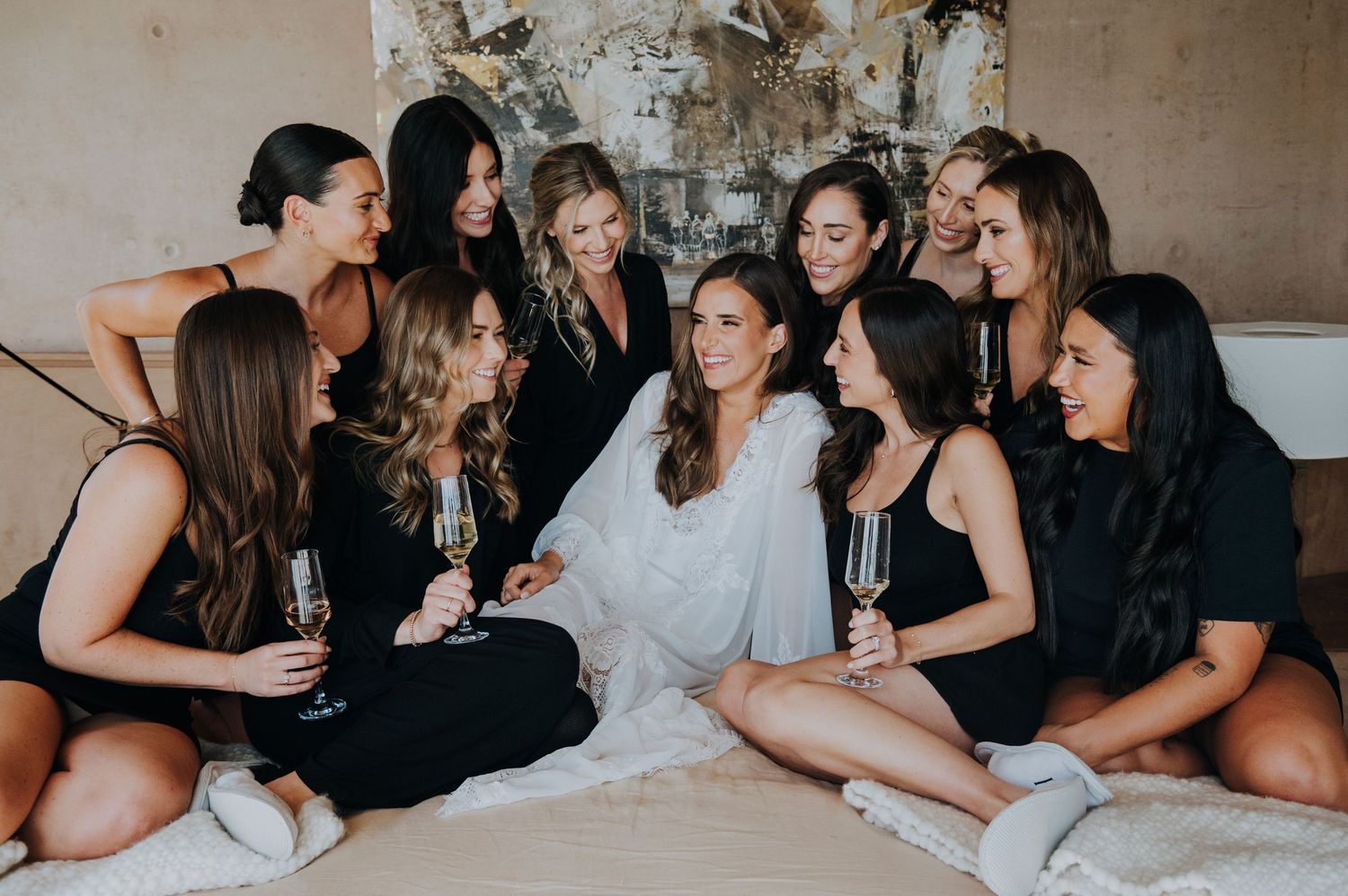 Bride in white robe surrounded by bridesmaids in black, toasting with champagne glasses on a bed.