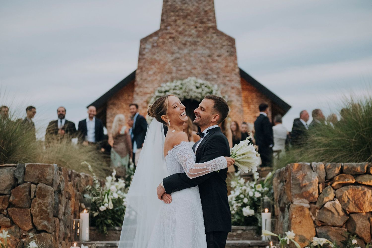 Bride and groom share a joyful moment at an outdoor wedding ceremony beside a rustic stone chapel.