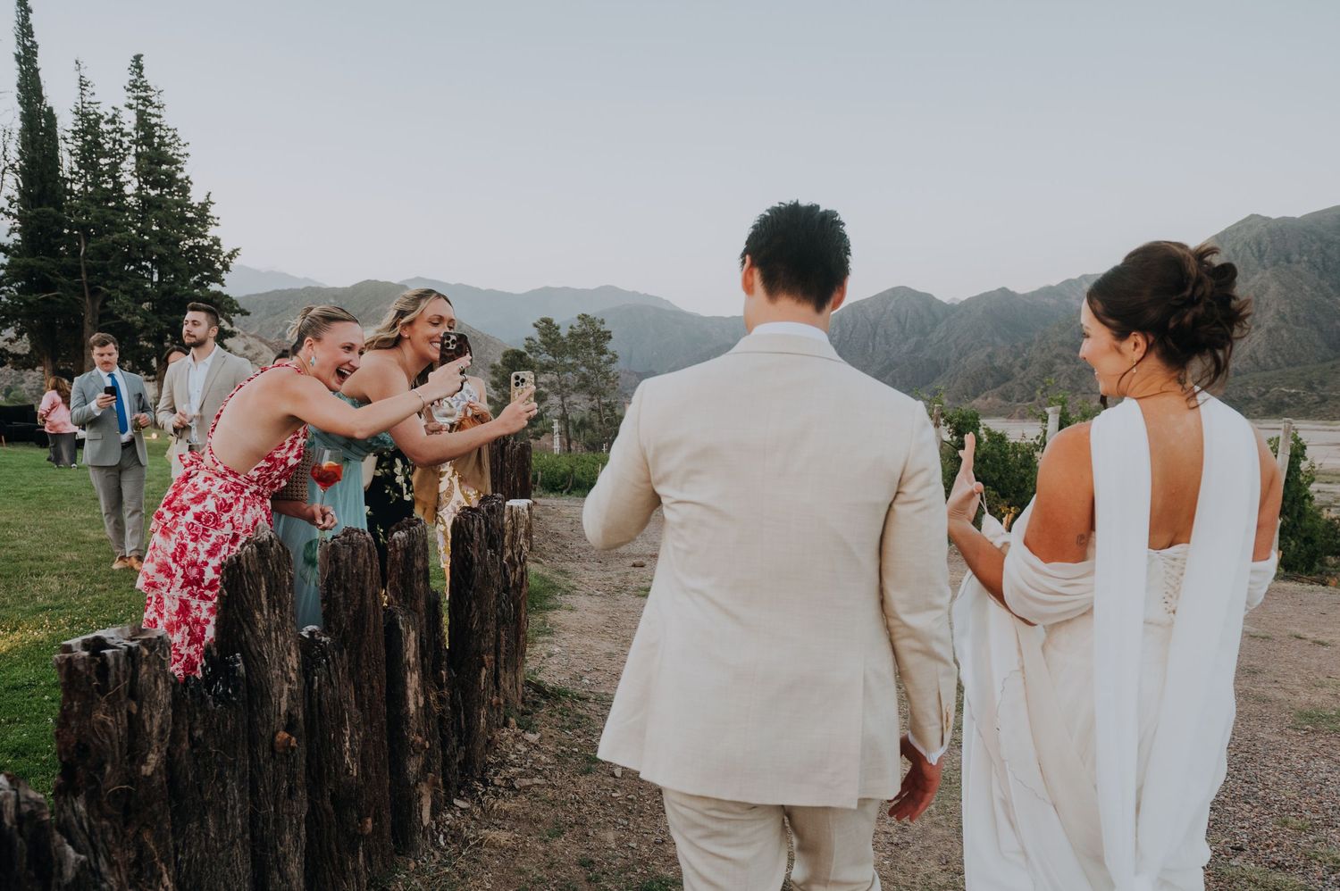 Wedding guests cheering as bride and groom walk past wooden posts at outdoor mountain venue.