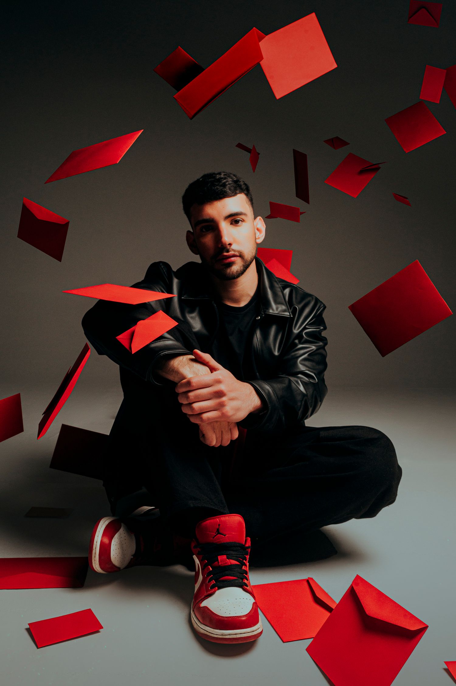 LETTER Young man in black leather jacket and red sneakers sits on floor surrounded by floating red envelopes.