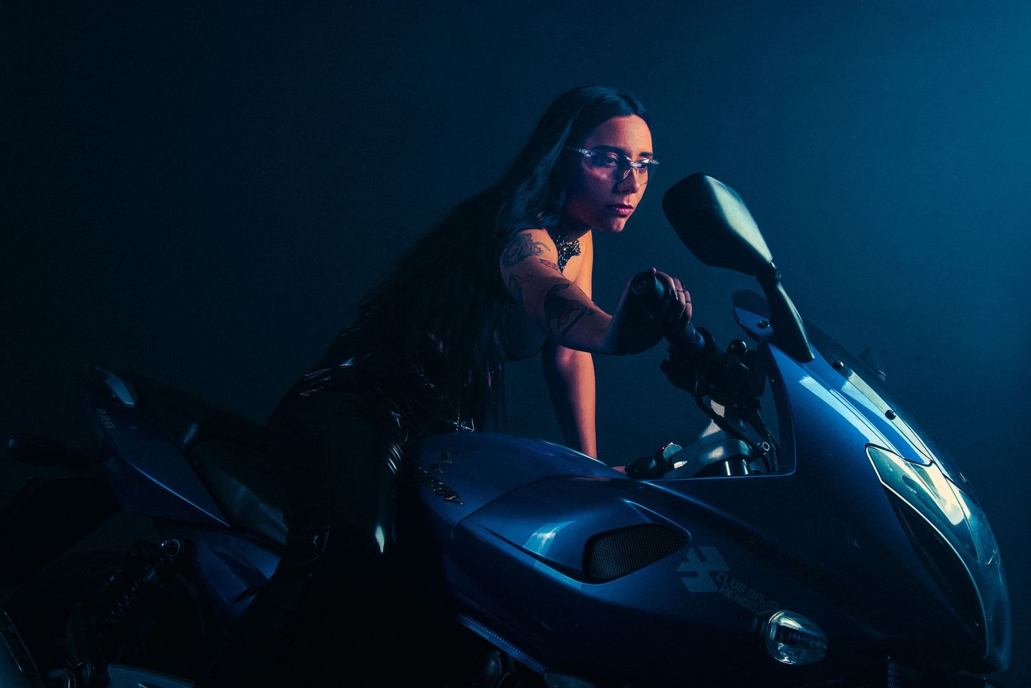 eve calleti Woman in dark outfit leaning on a motorcycle under blue studio lighting.