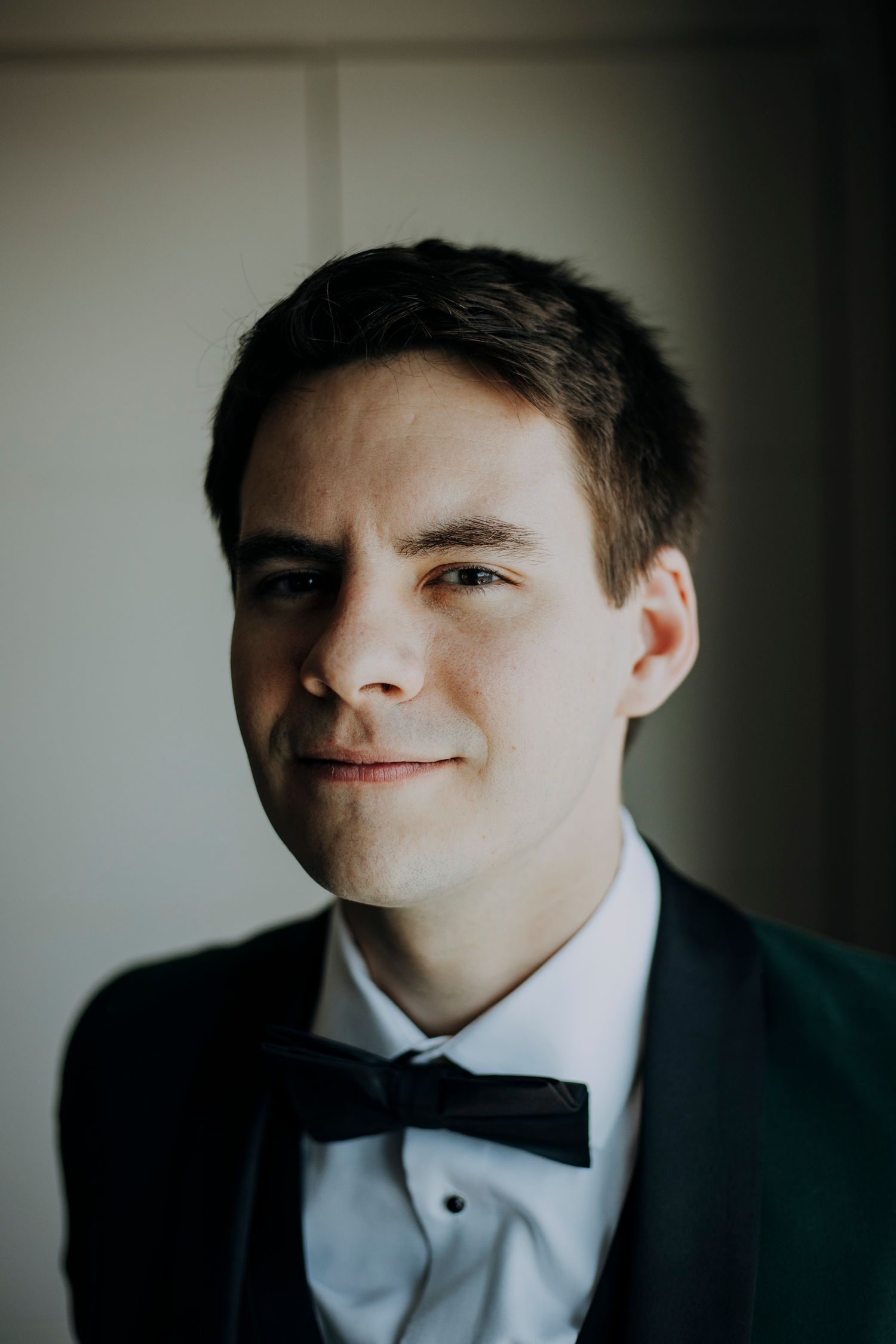 Young groom in black tuxedo and bow tie smiling softly at camera on wedding day.