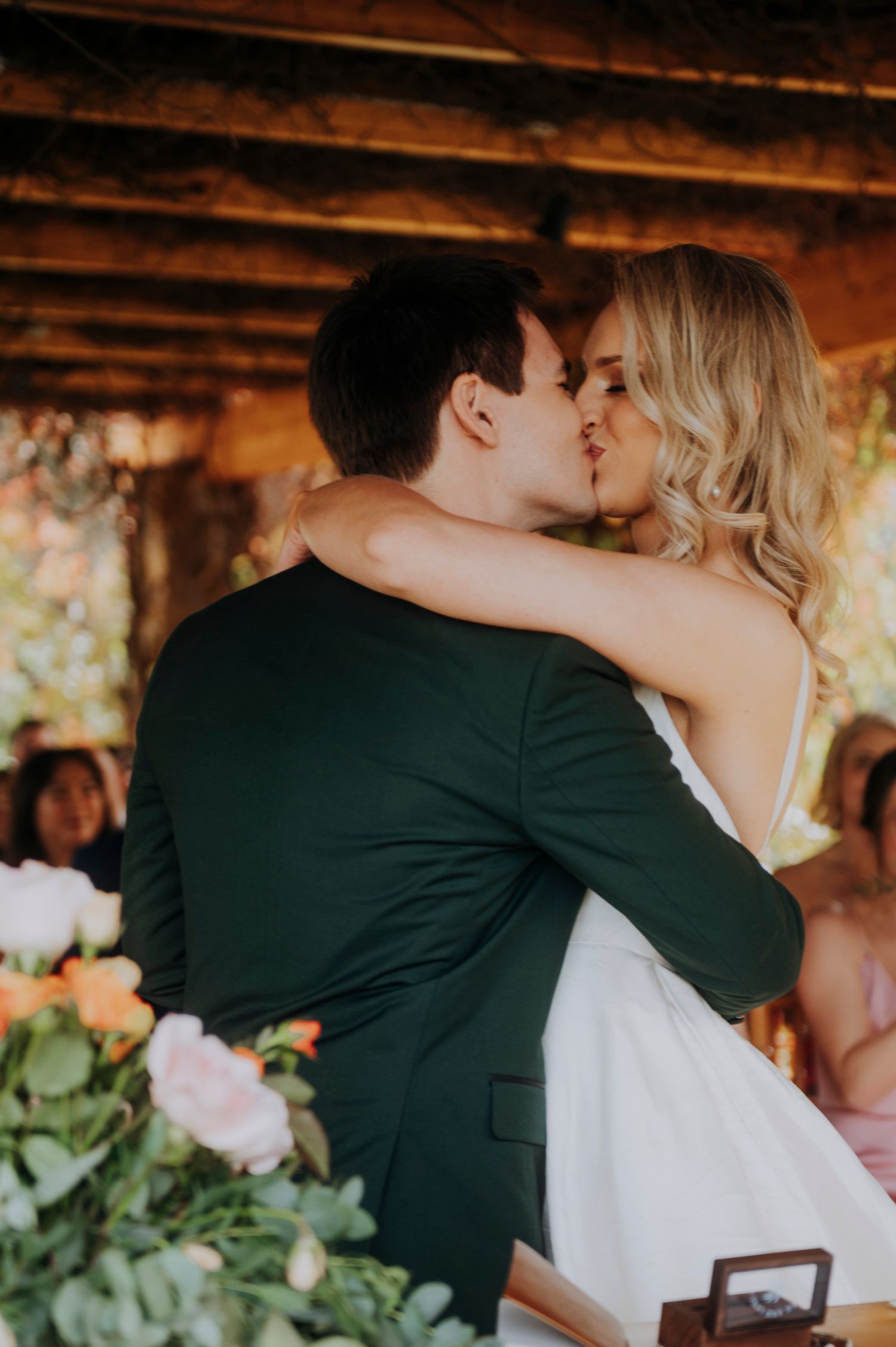 A couple shares their first kiss as newlyweds at a rustic wedding ceremony surrounded by flowers.
