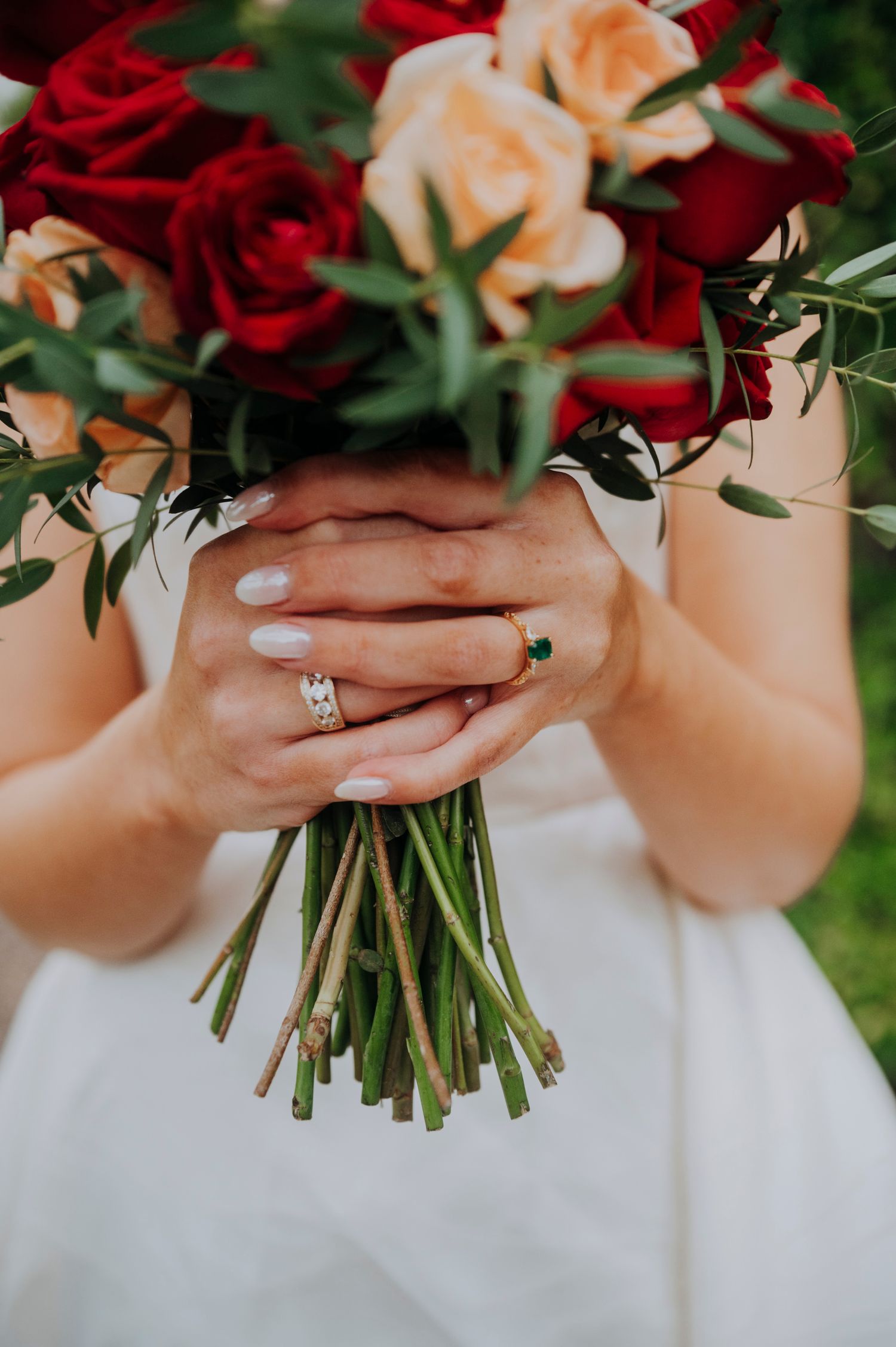 Bride holds red and peach rose bouquet showing diamond wedding band and emerald engagement ring.