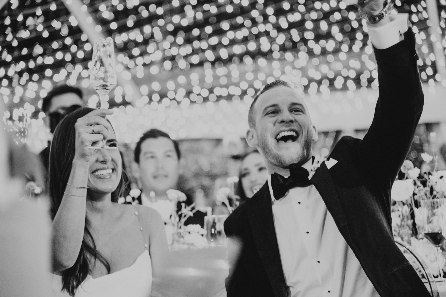Black and white photo of bride and groom celebrating joyfully under twinkling fairy lights at reception.