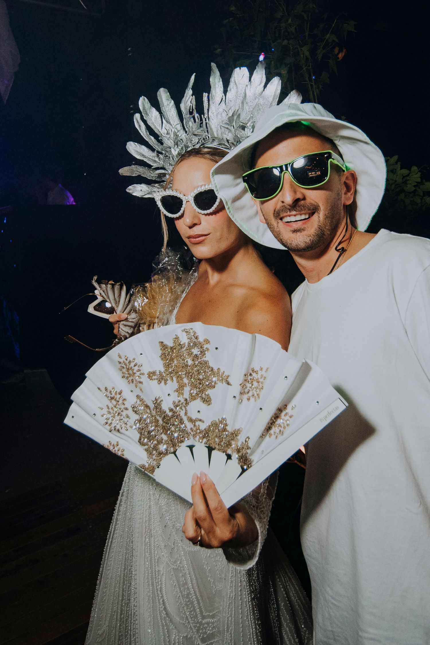 Two people in white costumes with sunglasses and silver headdress pose at a party, holding a decorative fan.