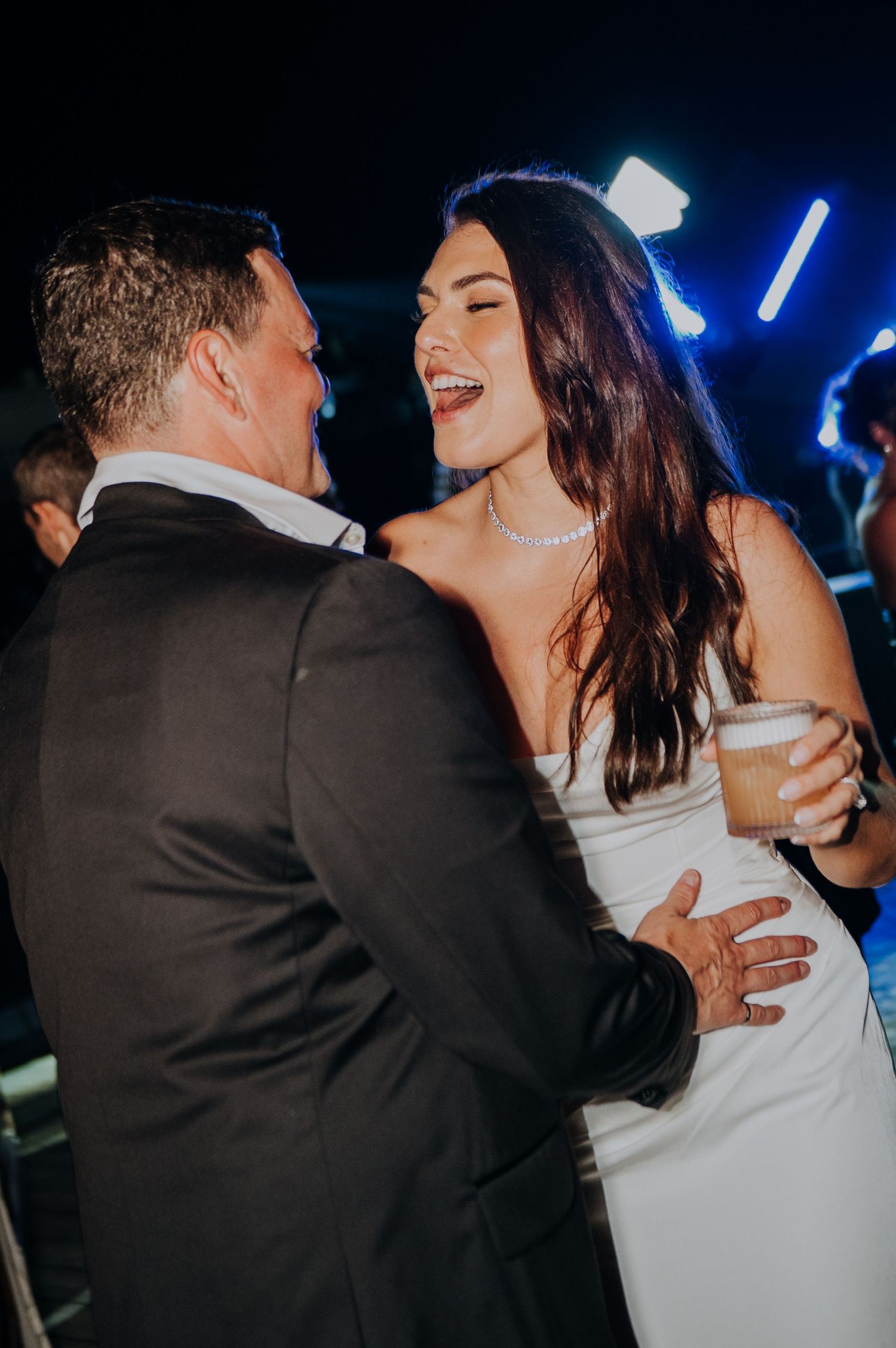 Joyful bride in white dress laughing while dancing with groom at nighttime wedding reception.