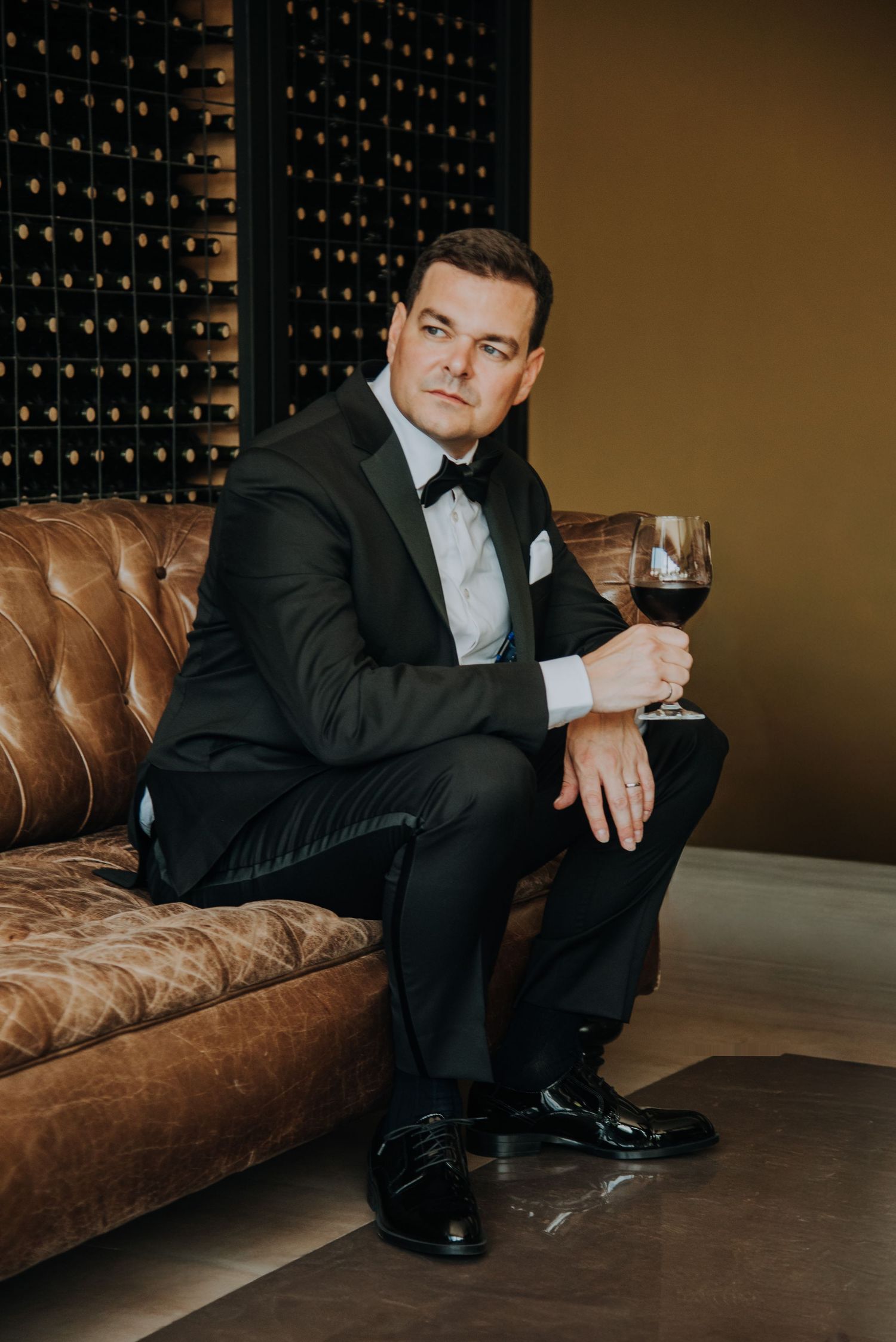 Elegant groom in black tuxedo sitting on leather sofa holding red wine glass in front of wine cellar.