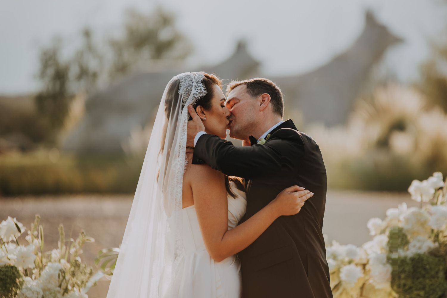 Newlyweds sharing a romantic kiss outdoors with floral arrangements and soft sky backdrop.