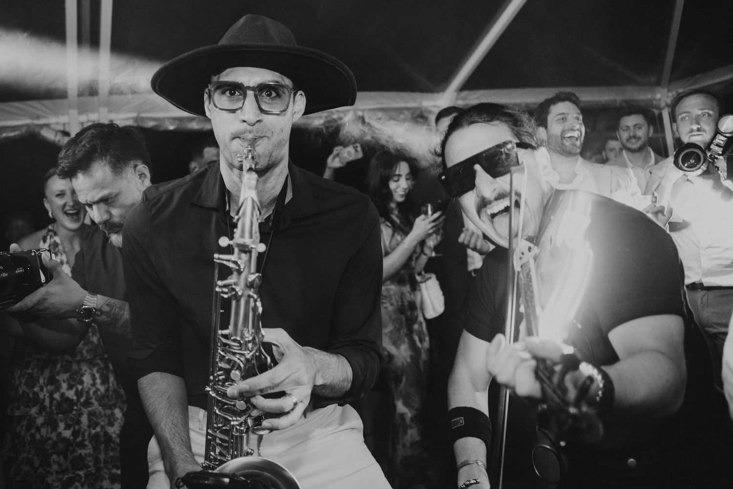 Musicians playing saxophone and trumpet at a lively outdoor event in black and white photo.