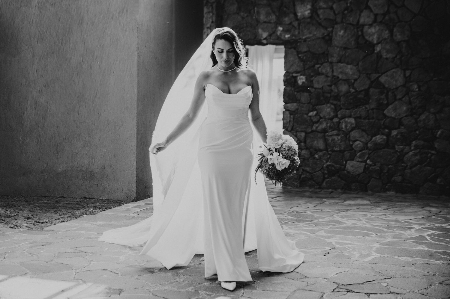 Bride in white strapless gown and veil holds bouquet in black and white wedding photo at stone venue.