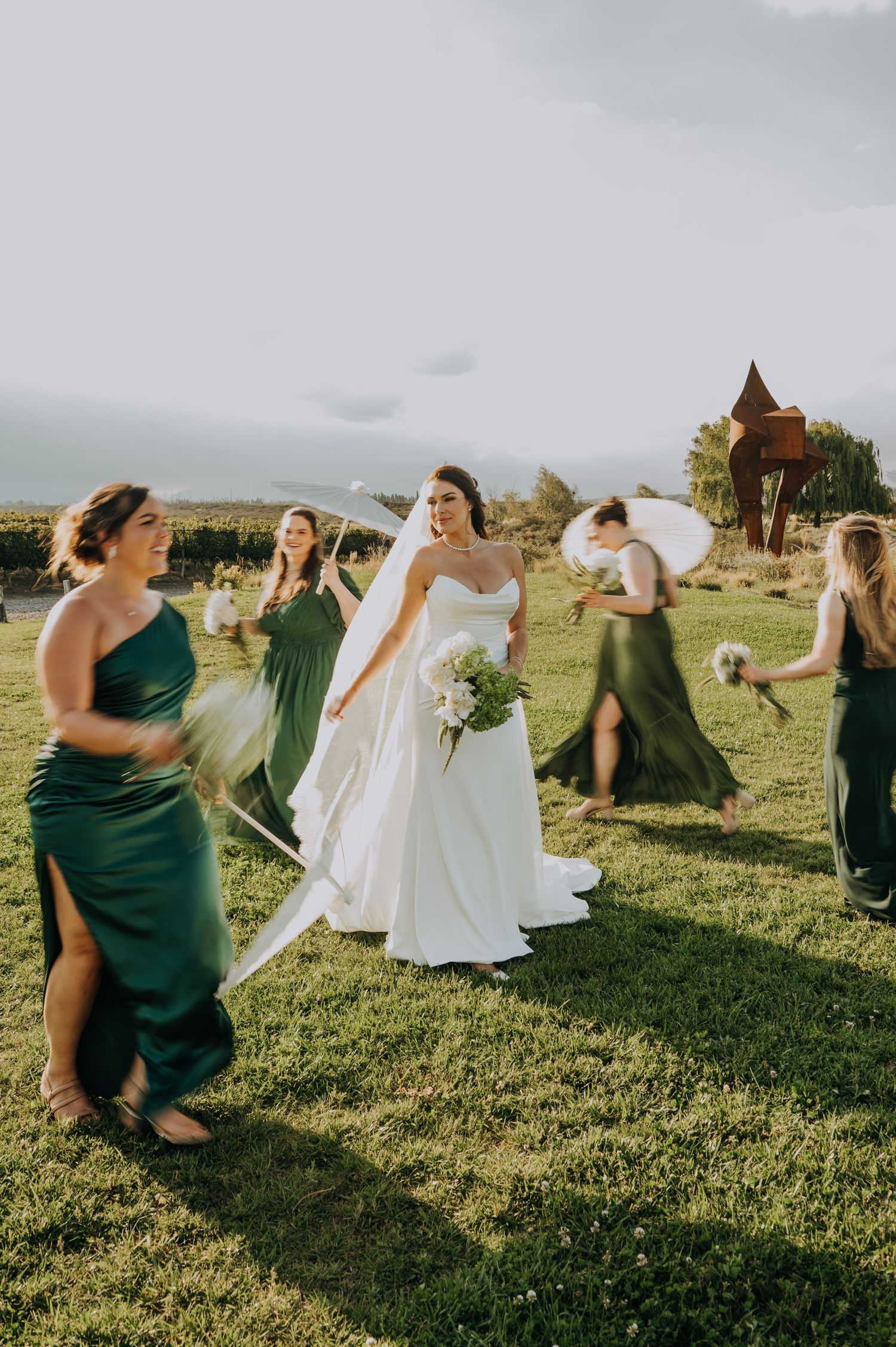 Bride in white gown walks with bridesmaids in emerald green dresses across a lush green lawn at outdoor wedding.