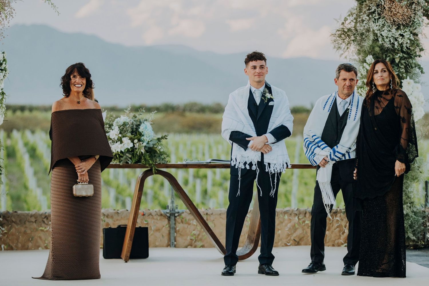 Jewish wedding ceremony outdoors with groom in tallit, officiant in brown dress, and family against vineyard backdrop.