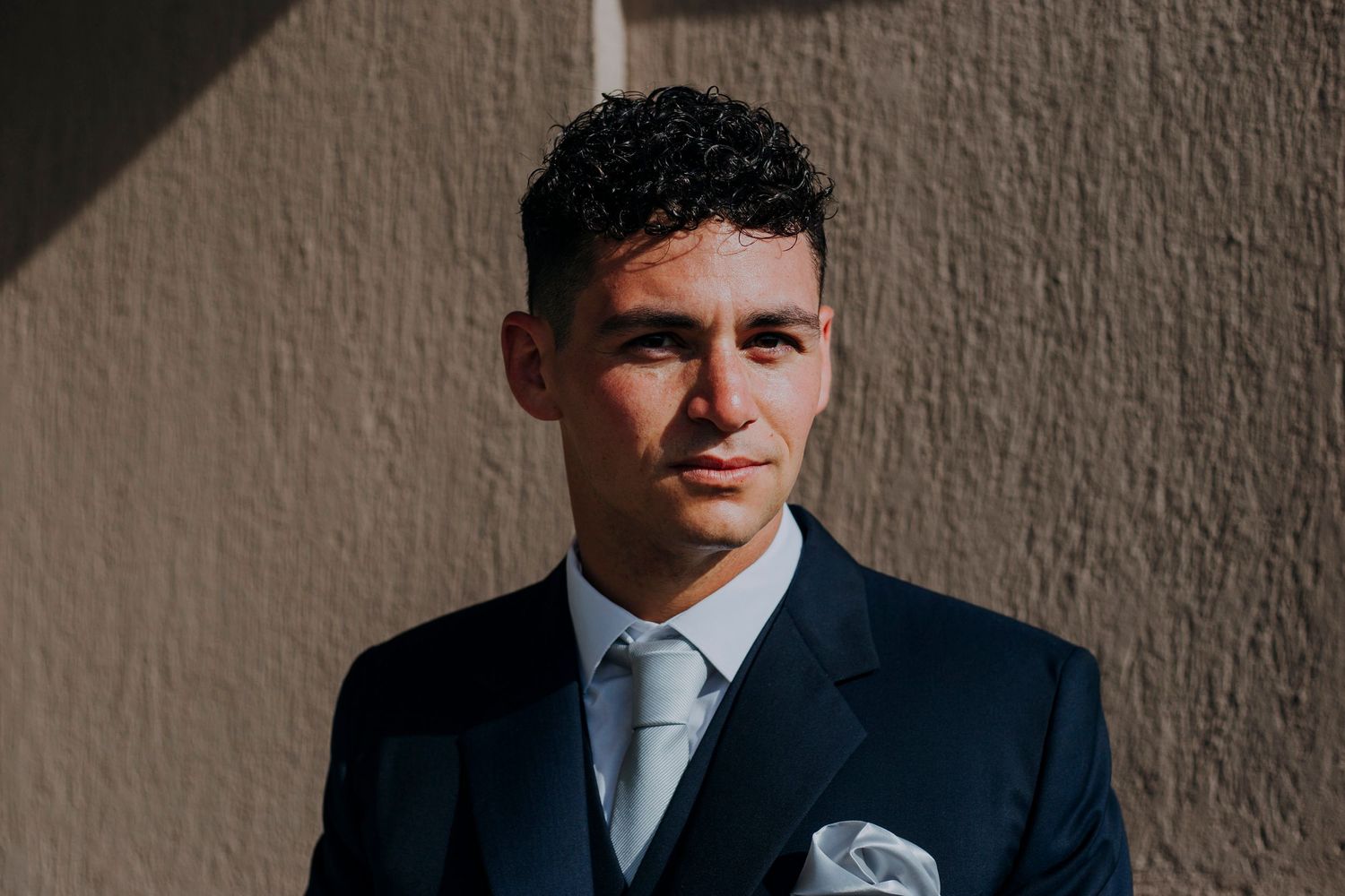 Handsome groom in navy suit and grey tie posing against sunlit wall before wedding.