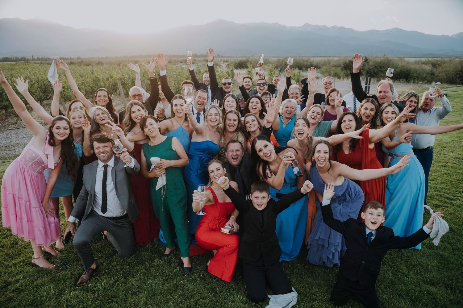 Large joyful wedding party group photo outdoors with mountain backdrop, guests cheering.