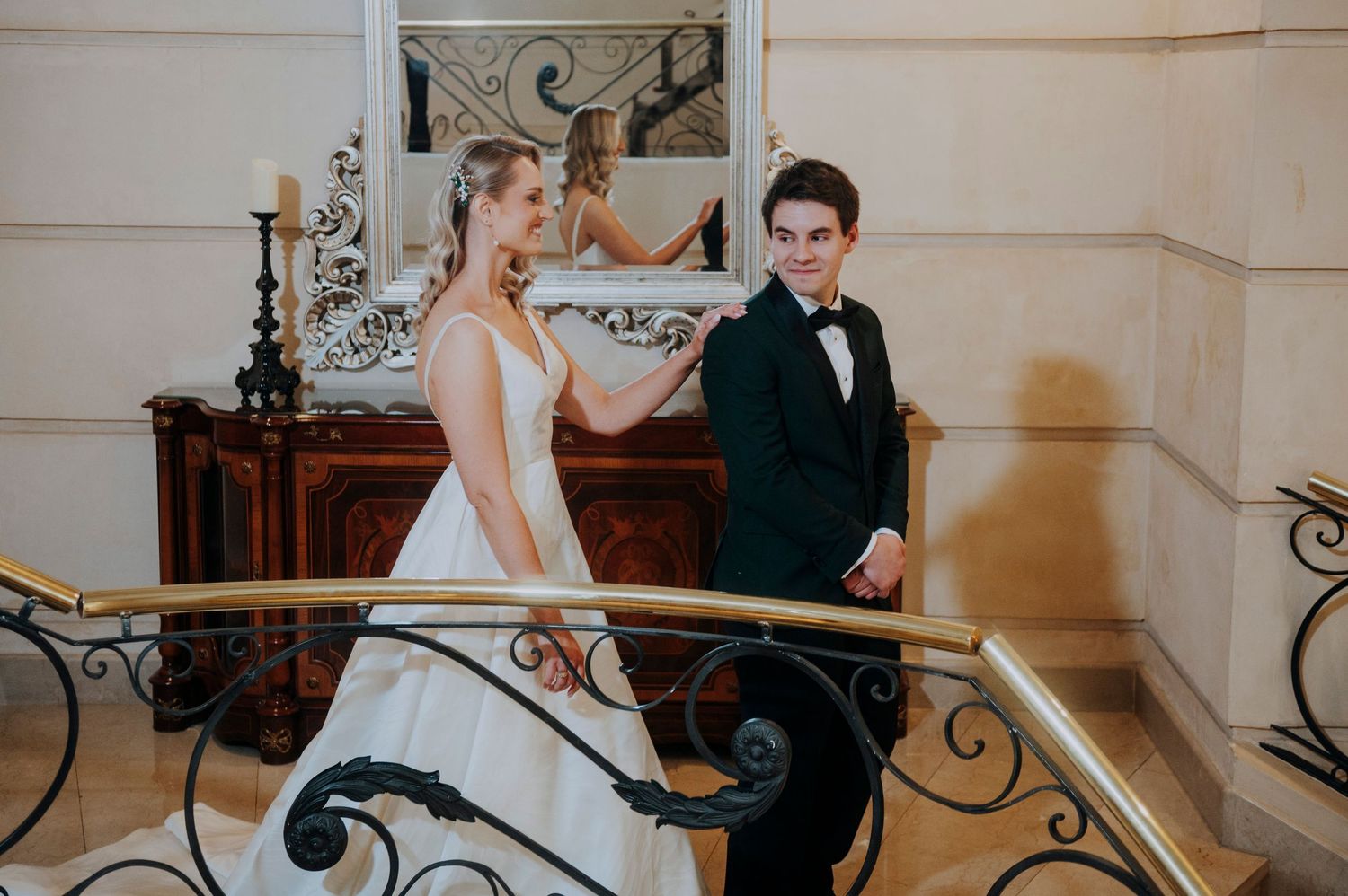 Bride and groom standing on an elegant staircase with gold railing in a grand wedding venue.