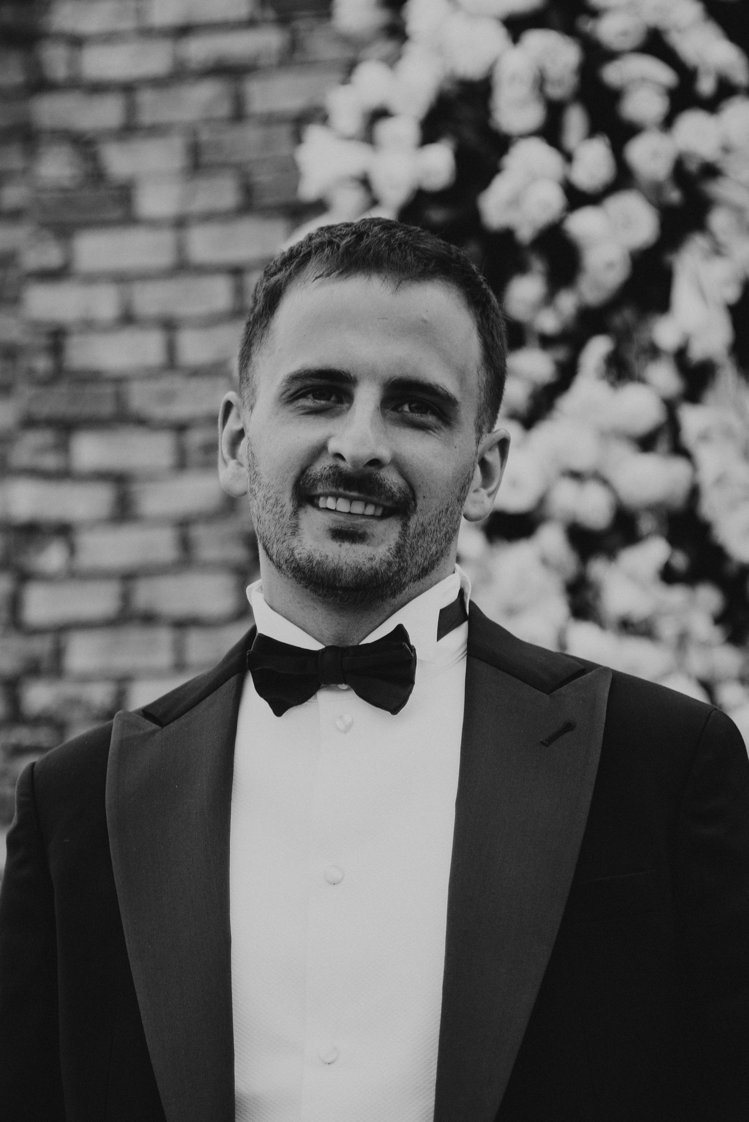 Groom in black tuxedo with bow tie smiling at wedding ceremony with floral backdrop.
