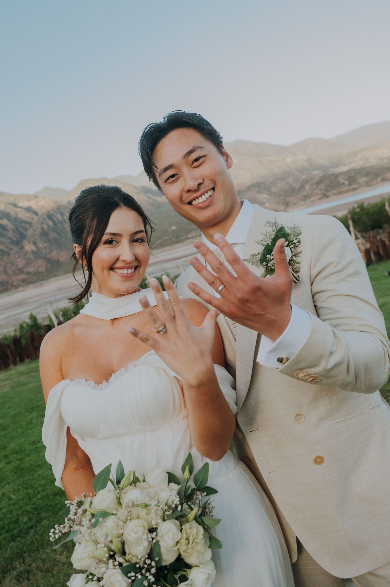 Newlyweds smiling and showing wedding rings with mountain landscape backdrop on their wedding day.